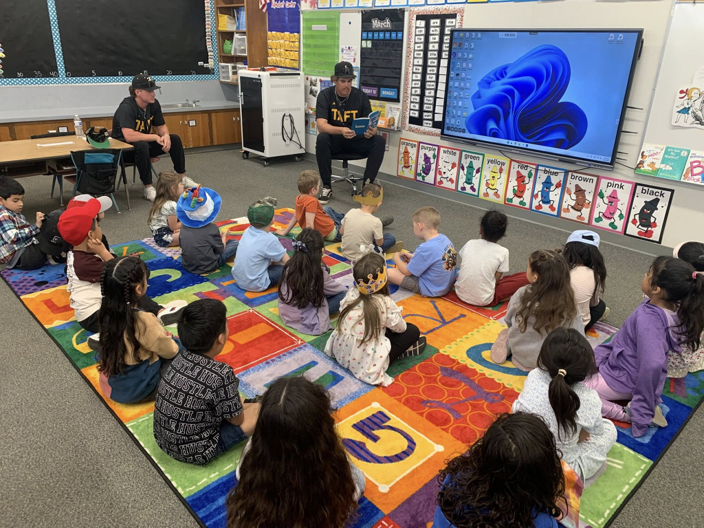 Two seated college athletes read books to 22 elementary school children in a classroom. 